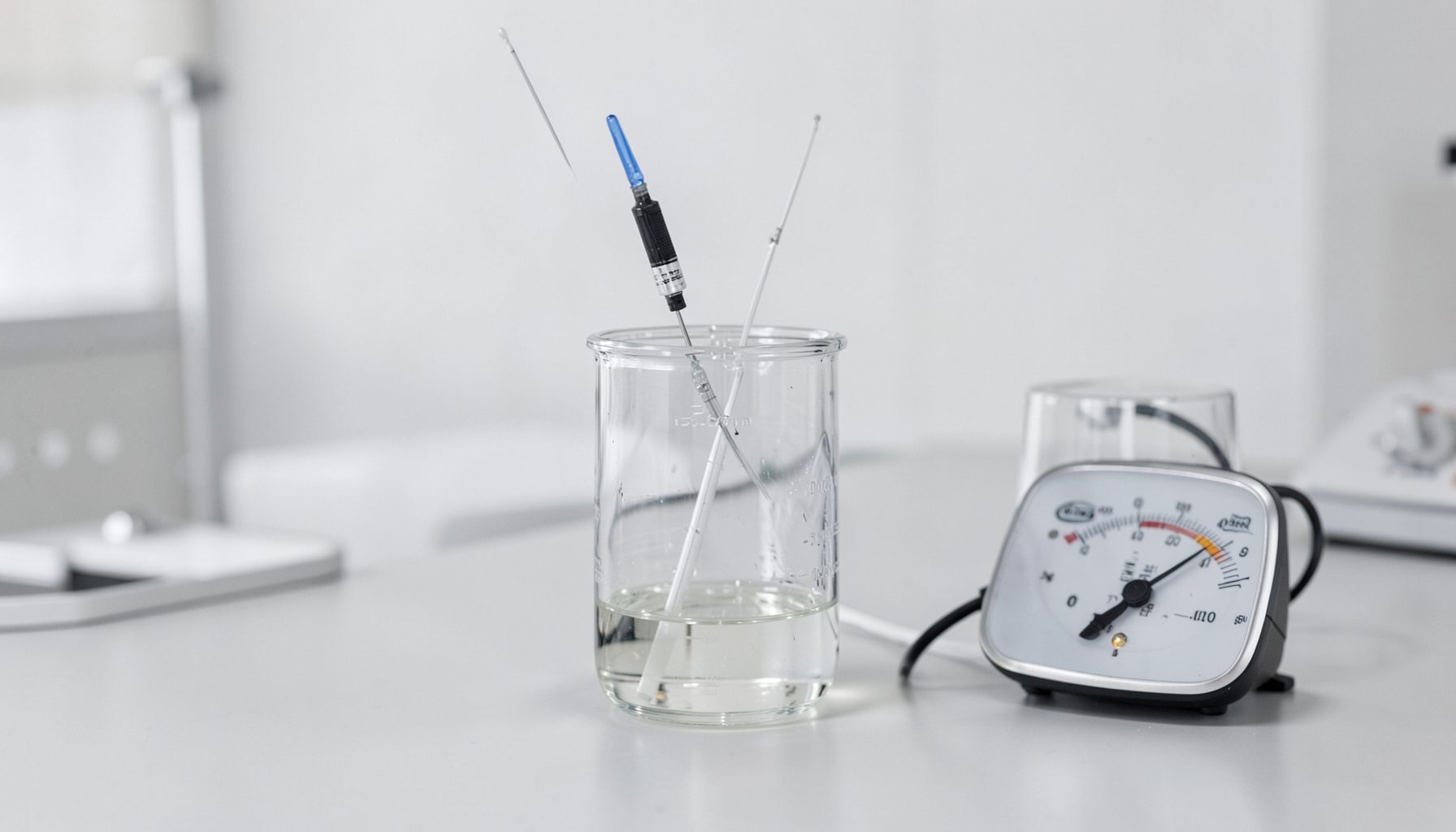 A laboratory setup for potentiometric measurement showing a reference electrode and an indicator electrode immersed in a beaker of solution, connected to a voltmeter. The scene has a clean, academic feel with neutral gray and white tones.